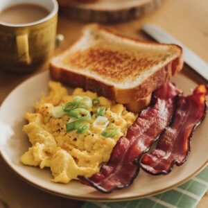 "Delicious bacon and egg breakfast with crispy bacon strips and sunny-side-up eggs on a white plate, accompanied by toast and a sprinkle of herbs."