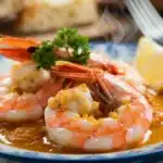 A close-up image of a shrimp bathed in a golden garlic butter sauce. The shrimp is glistening, and steam is rising from the sauce.