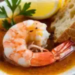 A close-up image of a shrimp bathed in a golden garlic butter sauce. The shrimp is glistening, and steam is rising from the sauce.