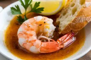 A close-up image of a shrimp bathed in a golden garlic butter sauce. The shrimp is glistening, and steam is rising from the sauce.